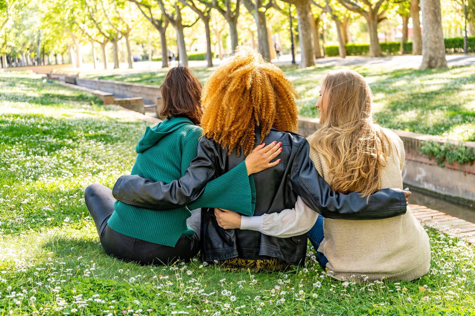 Three young friends, each from a different background, showcase the strength of their friendship as they pose in a back-to-camera embrace on a beautiful day in the park.